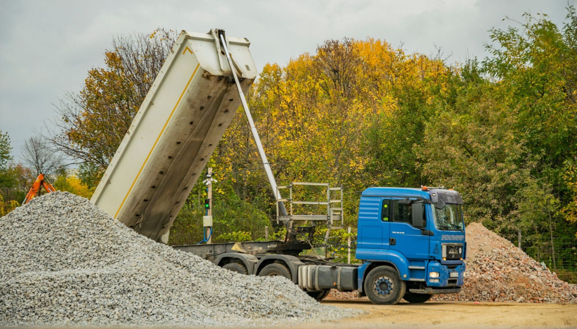 Blue dump truck unloading gravel at an outdoor construction site surrounded by autumn trees.